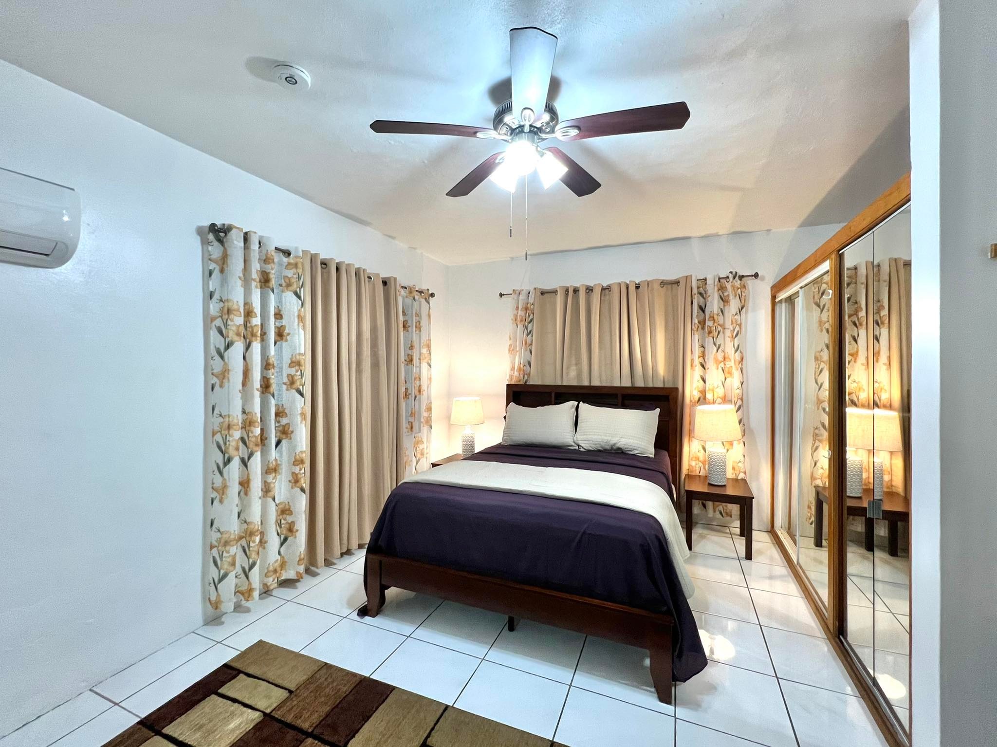 322141629_888080145567702_330212582415547576_n Living Room with 2 basket lamps hanging from the ceiling, grey couch against a grey wall, orange ottoman, circular marble tea table, 2 ornament floor plants, brown lazy chair to the right, wide western run under everything, and deep hallway to the far right, white flowy drapes to the far left.